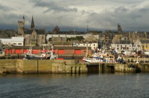 A harbour in Scotland, with a series of grey buildings and church spires in the background, and the sea wall with a selection of white fishing boats at anchor in the foreground.