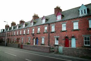 A suburban street view with a terrace of red brick houses. Each terraced house has a ground floor window and door, a pair of first floor windows, and a dormer window in the roof.