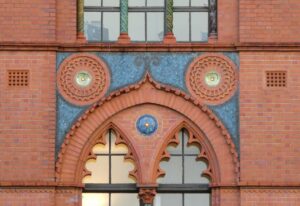 A close-up of the side of a historic red brick building with very ornate features. There is a blue tiled mosaic and circular motif above a window, which is a pointed shape with round edges.
