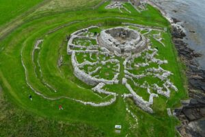 An aerial photograph of a ruined broch structure next to the sea. The series of concentric walls are made of grey stone on a grass slope. There are people walking around the structure.