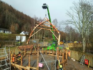 A very large wishbone-shaped pair of wooden beams being lifted by a crane. People in protective workwear are guiding the cruck, which looks like an upside down V, into place