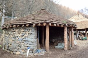 A traditionally constructed roundhouse building, with another in the background. This one has a turf roof, with the outer wall made of dry stones. The entrance is held up by a row of straight tree trunks.