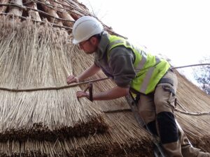 A person standing on a ladder working on a thatched roof of a traditional roundhouse building. They are wearing a high visibility jacket and a protective hard hat and are pushing a series of dry reeds into place.