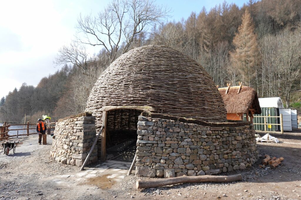 A woven wood roundhouse with a large open doorway. There is a head-height stone wall running around the outside as if enclosing it, with other traditional buildings in the background.