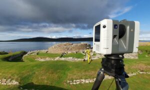 A digital surveying camera mounted on a tripod overlooking the remains of a historic circular stone broch next to the sea.