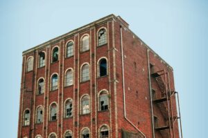 The top four storeys of an abandoned brick factory building. Each floor has six windows, many of which have broken or missing glass, and there is a metal fire escape on the other side.