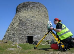 A person wearing a high visibility jacket kneels down in front of a piece of digital recording equipment on a tripod, and looks through a viewfinder at an ancient broch tower; a chimney shaped stone building against a blue sky.