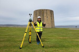 A person wearing a high visibility jacket and baseball cap stands next to a surveying camera on a tripod set in front of a two storey round tower. They are smiling and giving two thumbs up.