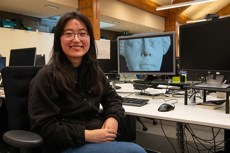 A person with long black hair and square framed glasses smiling whilst seated at a desk in an office. On one of the computer screen's behind them, the background is a 3D model of Mary Queen of Scot's death mask