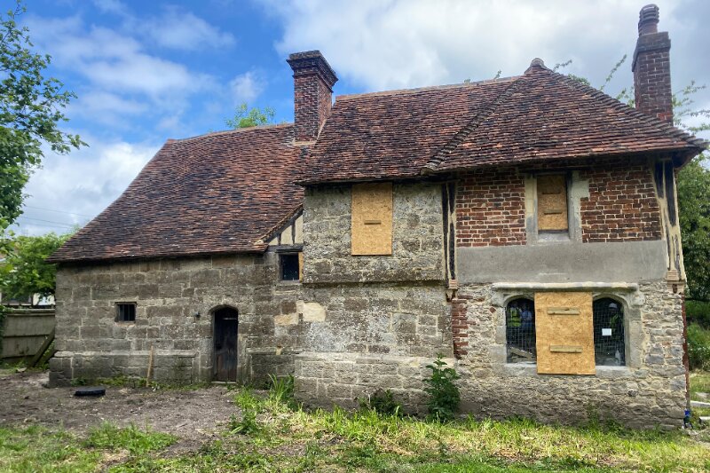 A very old stone and brick building, with a tiled roof and two chimneys. The building is many different colours and masonry styles, and looks derelict as it has boarded up windows.