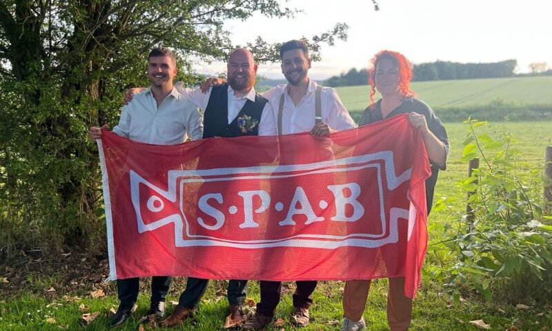 Four people standing in a field holding a large red banner that says SPAB. They are all wearing wedding clothing and are smiling.