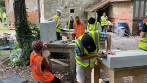 A group of people in high-visibility vests, working with hand tools on different projects in the grounds of an old brick and stone building.