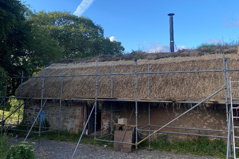 A very old looking single storey building with a new looking thin metal chimney. The roof is thatch and the walls are covered in scaffolding.