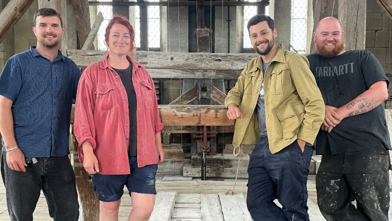 Four people facing the camera and smiling, standing inside an old wooden belfry. They are leaning on a wooden crossbeam.