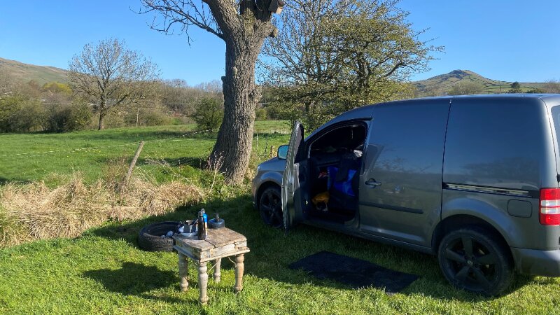 A grey van parked in a grassy field with the door open. Next to the van is a table with cooking items on it.