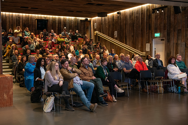 Many people seating in rows inside a wood-clad auditorium, listening to a speaker.