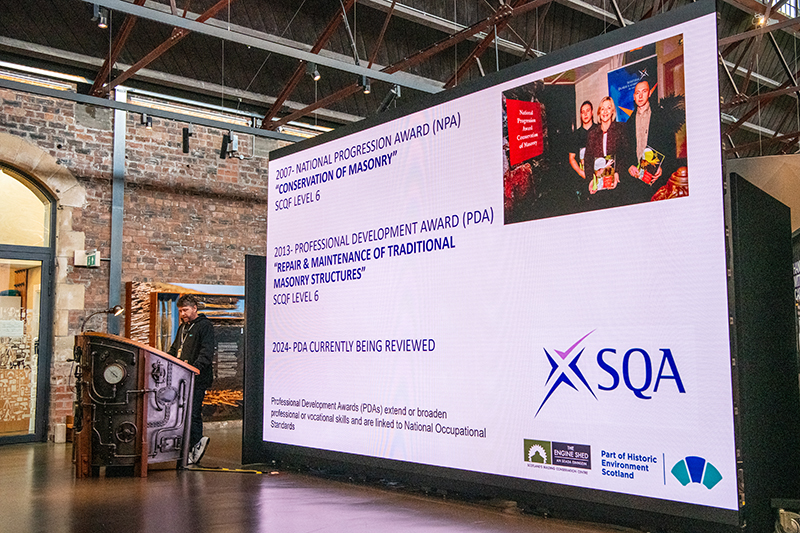 A very large, free-standing screen inside the Engine Shed main space. In front of the screen, someone is standing speaking at a large lectern.