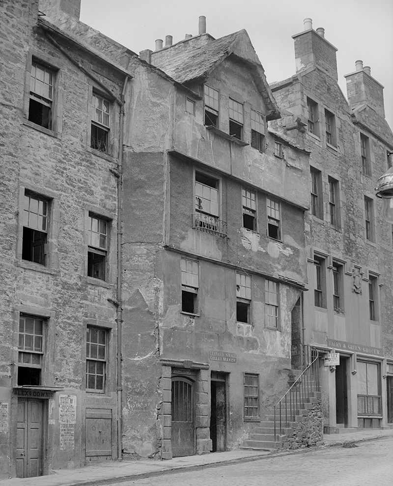 A row of old, sandstone tenements with three storeys. There are many sash and case windows, wooden doors, slate roofs and chimney stacks.