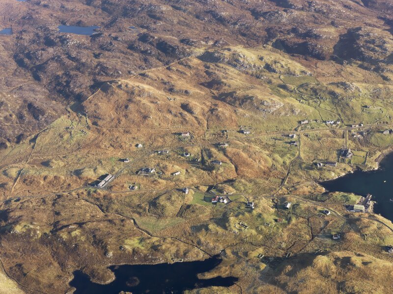 An aerial view from very high up of a collection of scattered buildings in a remote island location. There are hills in the background, with about thirty detached buildings spread across the landscape, and inlets of the sea in the foreground.