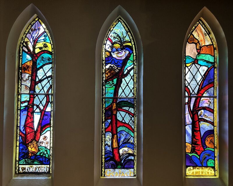 Three tall, narrow stained glass windows against a dark background. Each one features a tree against a colourful background.