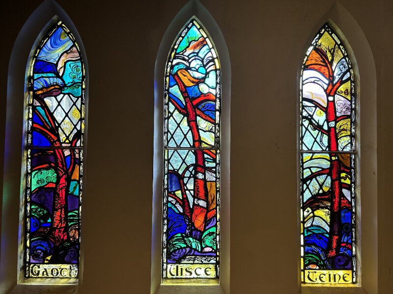 Three tall, narrow stained glass windows against a dark background. Each one features a tree against a colourful background.