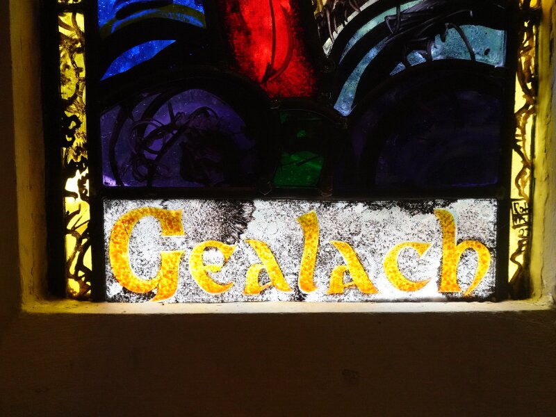 Close-up detail of the bottom half of a historic stained glass window. The colourful glass is bordered by thick black lines, and the Gaelic word 'Gealach' at the base.