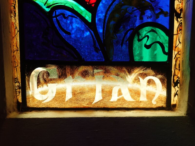 Close-up detail of the bottom half of a historic stained glass window. The colourful glass is bordered by thick black lines, and the Gaelic word 'Grian' at the base.