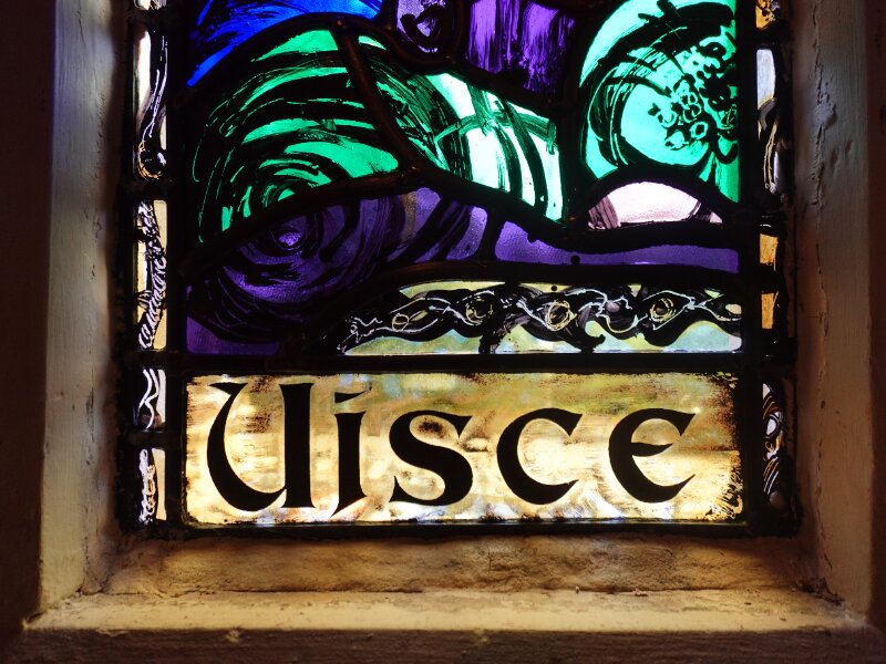 Close-up detail of the bottom half of a historic stained glass window. The colourful glass is bordered by thick black lines, and the Gaelic word 'Uisce' at the base.