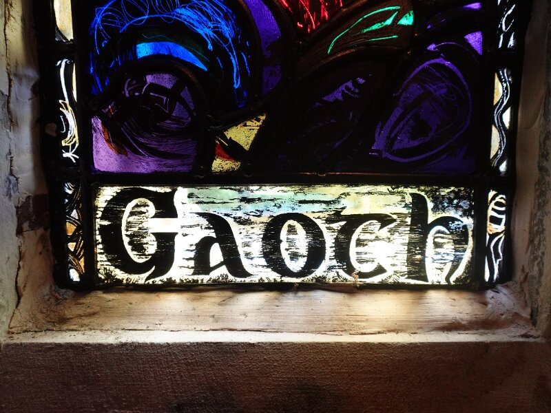 Close-up detail of the bottom half of a historic stained glass window. The colourful glass is bordered by thick black lines, and the Gaelic word 'Gaoth' at the base.