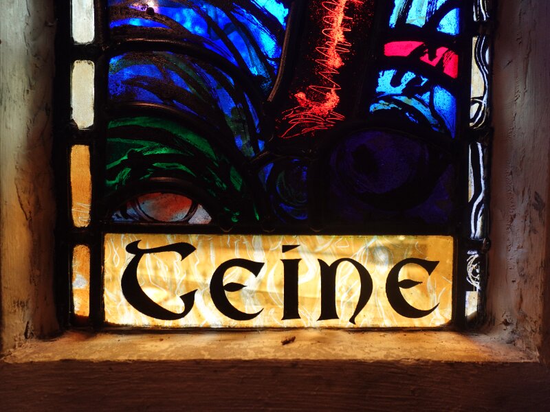 Close-up detail of the bottom half of a historic stained glass window. The colourful glass is bordered by thick black lines, and the Gaelic word 'Teine' at the base.