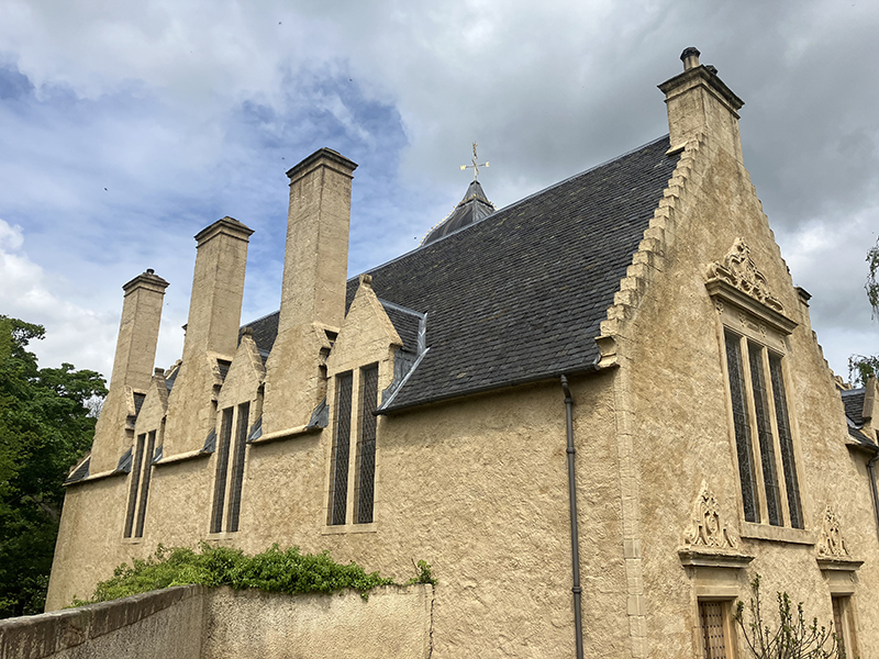 A historic building with a steep slate roof, large chimney stacks and narrow but tall windows. The building is covered in a muted yellow limewash.