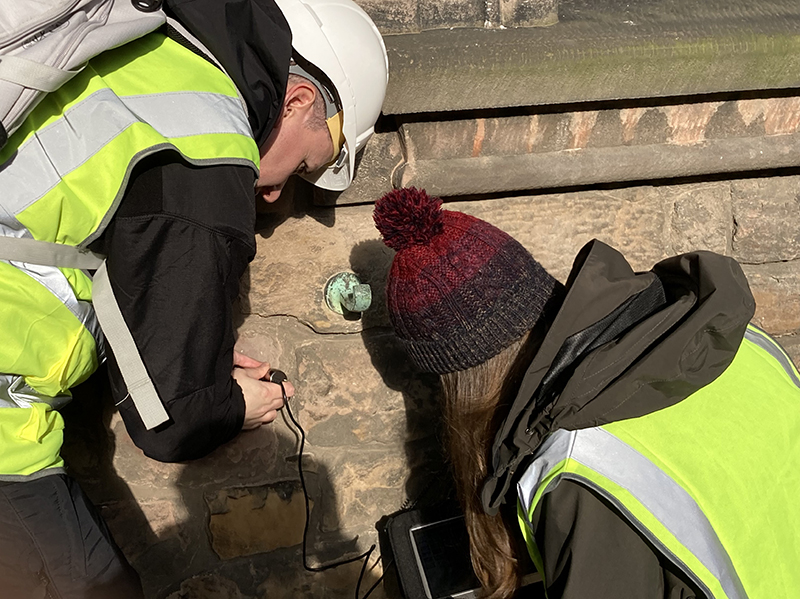 One person is holding a piece of equipment up to a stone building and the other is looking at the monitoring screen attached to the device.