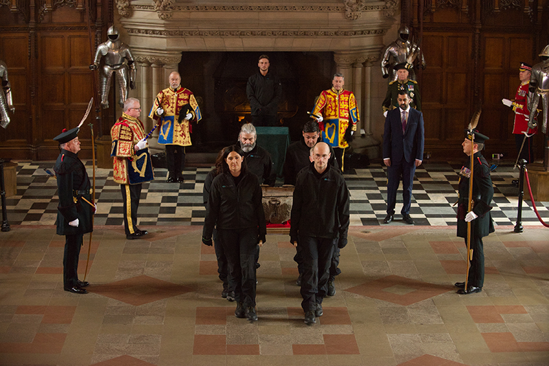 A group of people, standing in a room with the stone of destiny. Some are wearing official Scottish and army uniforms, some area dressed in all black HES uniforms.