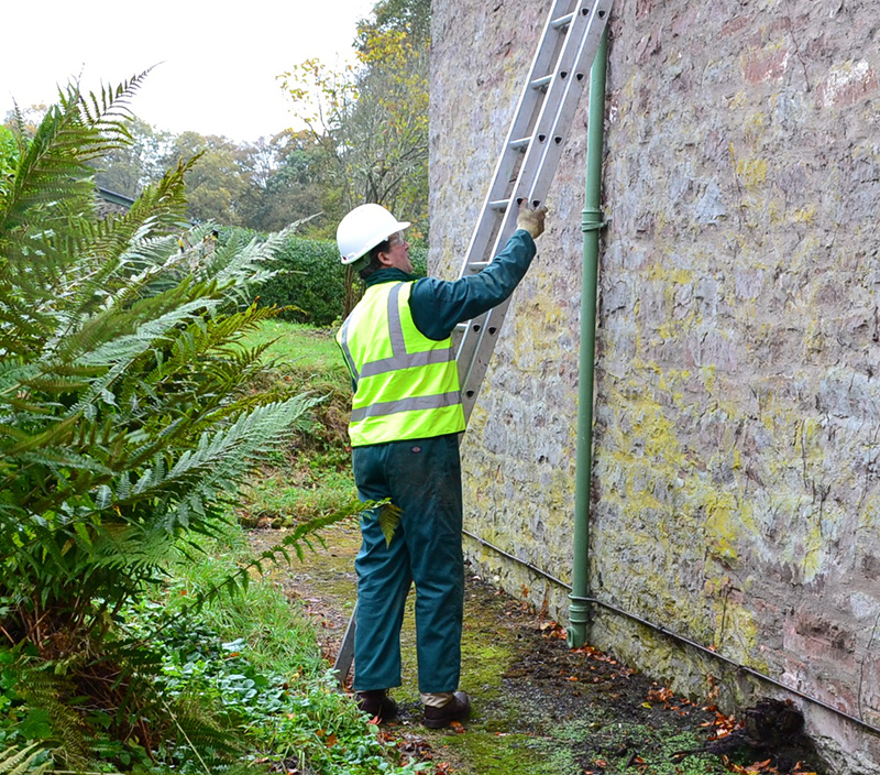 A person wearing overalls, a high visibility vest, a hard hat and gloves, holding a ladder up to the stone wall of a building.