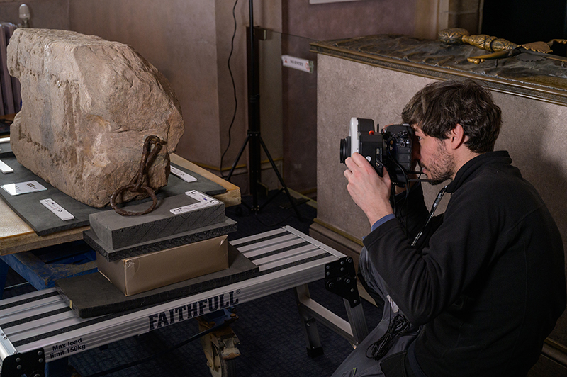 A person crouching down with a camera, taking a photo of the stone of destiny