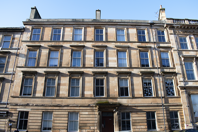 A row of blond sandstone tenements with four storeys and many rows of long sash and case windows.