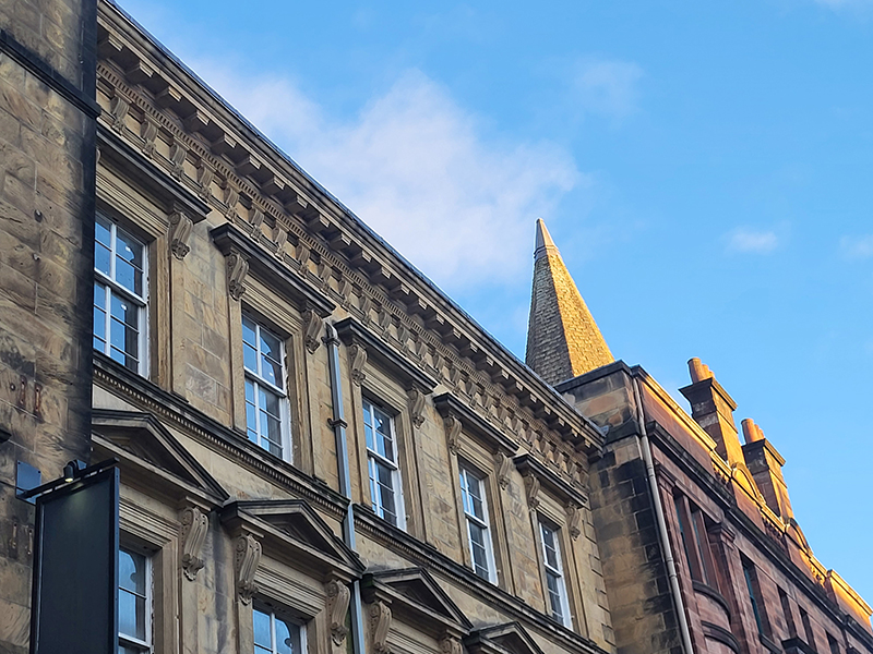 A row of traditional sandstone tenements with rows of sash and case windows and carve stone detailing above and around the windows.