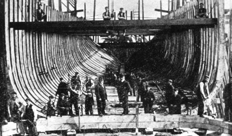 A group of shipbuilders standing inside the skeleton hull of a boat they're building