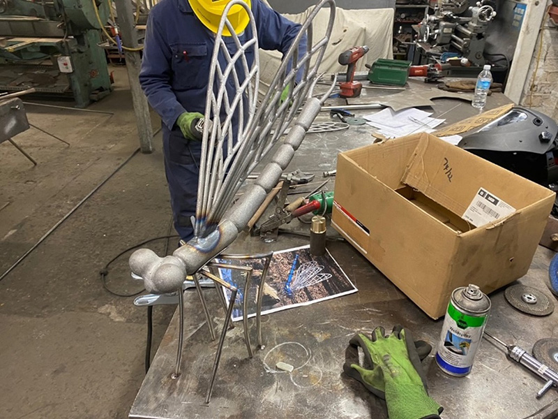 A large sculpture of a dragonfly made from metal. A person is working on it in a blacksmithing workshop.