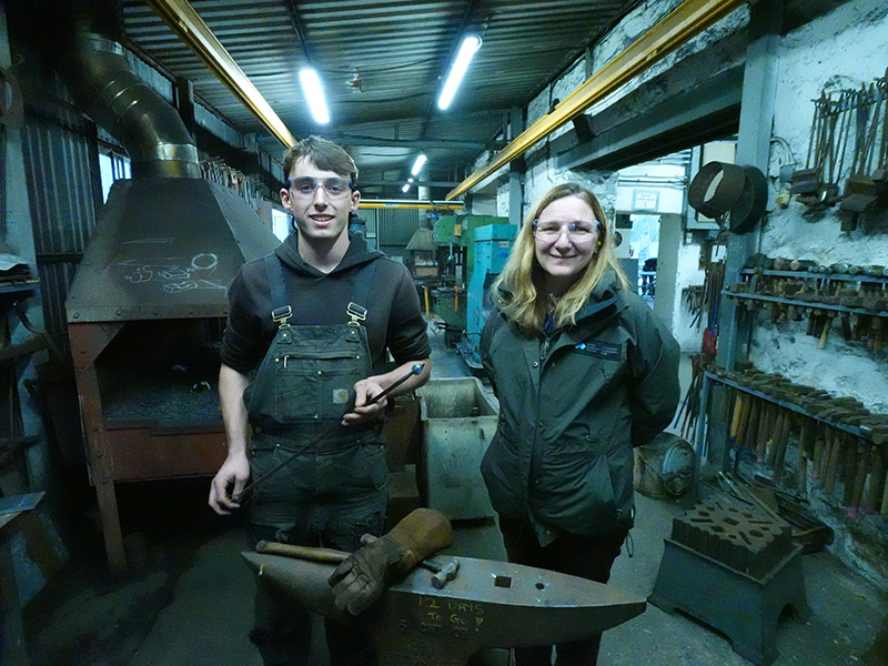 Two people standing behind an anvil in a blacksmithing workshop, wearing safety glasses. They are both smiling and of them is holding a piece of metal.