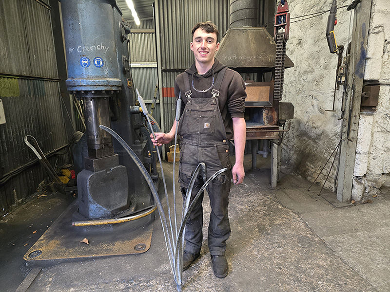 A person in a workshop wearing overalls, holding a metal sculpture of bulrushes, smiling.