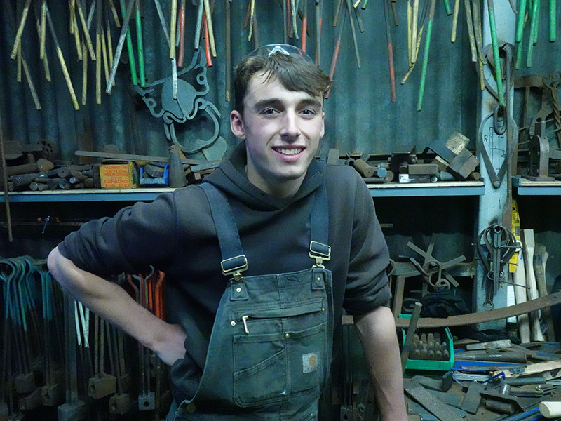 A person wearing work overalls standing in a workshop smiling with shelves covered in many tools behind them. They have short, dark hair and are wearing safety googles on top of their head.