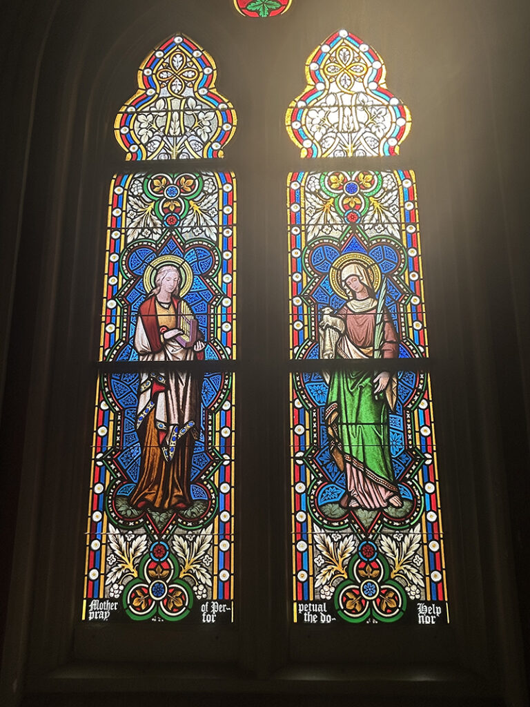 Two tall, thin stained glass windows in a church. Each one features a different Saint. The Saint on the left is holding a harp, while the Saint on the right is holding a lamb.