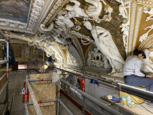 Scaffolding set up to ceiling height, with a person sitting on top of the scaffold, working on the ceiling. The ceiling has extremely ornate plasterwork on it, including sculptures of people and large swathes of gilding and other gold decoration.