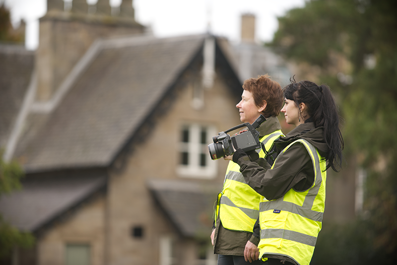 Two people wearing high vis vests standing outside a traditional stone building. One is holding a camera