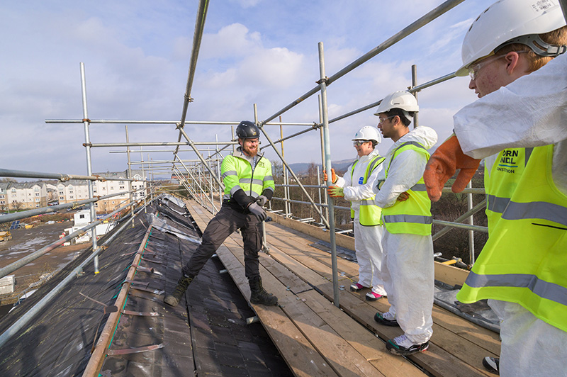 Three young people wearing overalls and hard hats, standing on top of scaffolding on a roof, speaking to a man in work gear.