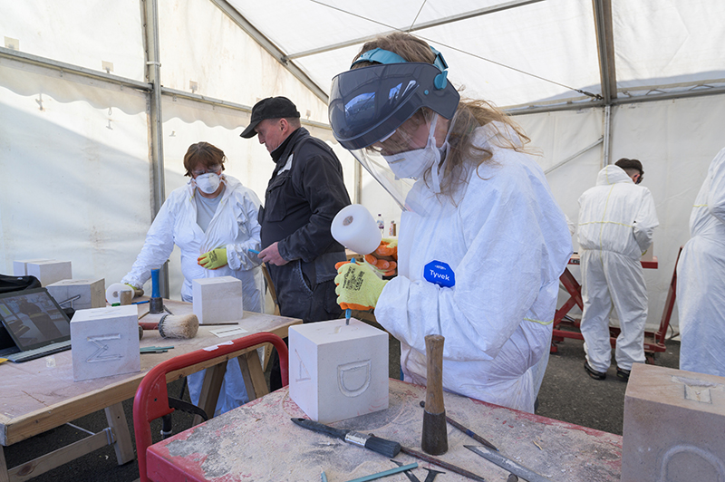 A person wearing overalls and a visor carving letters into a piece of stone in a marquée. There are other people in the marquée also carving into stone