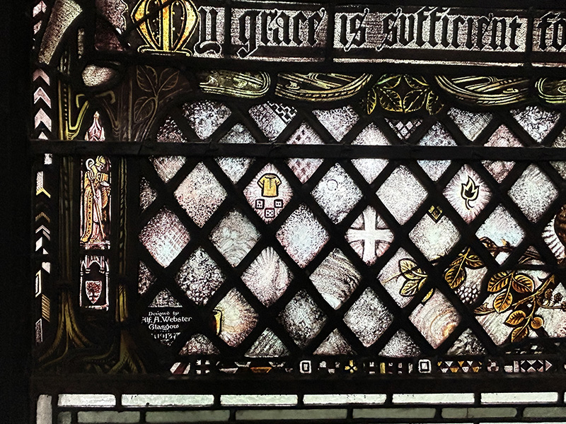 A stained glass window with criss crosses on it and different things within the diamond shapes like some dice, a t-shirt, a cross and a flower.