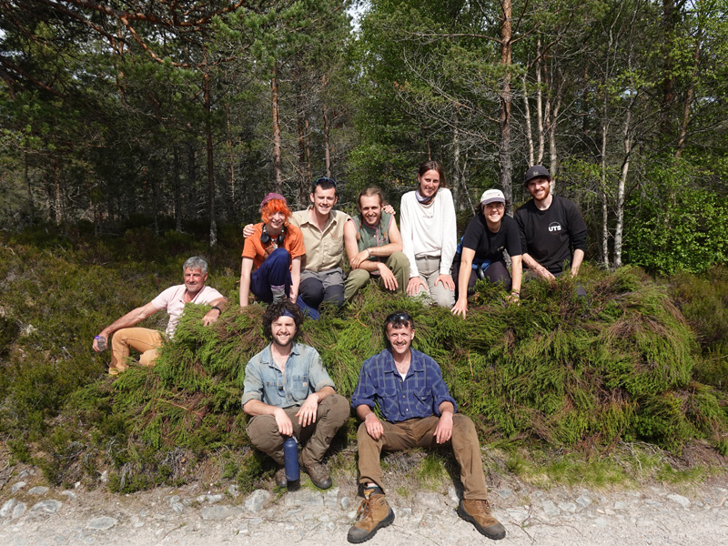 A group of 9 people, 7 of them are perched on top of a large pile of heather and two of them are sitting in front of the heather. They're all smiling.