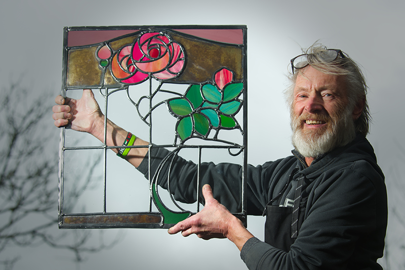 A person with glasses and a white beard holding up a beautiful stained glass window with a rose design
